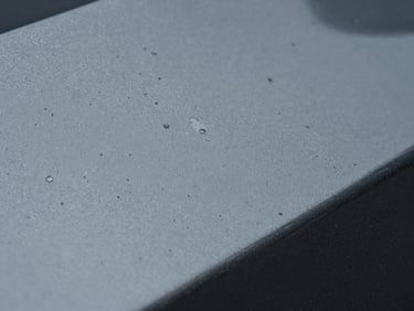 Extreme macro close-up of automotive paint surface under studio light, visible mineral deposits and water spot etching on dark metallic paint, overcast diffused lighting revealing micro-texture imperfections