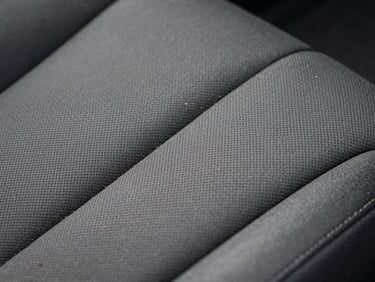 Close-up of automotive interior upholstery texture under controlled diffused lighting, fabric weave detail visible with embedded dirt particles before extraction treatment, high contrast revealing surface condition