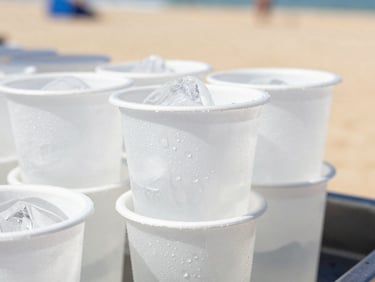 Macro close-up of pre-filled ice cups stacked on a beach club bar, bright high-key summer sunlight casting sharp refreshing shadows, condensation droplets