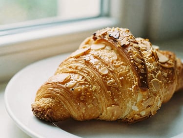 Close-up of a golden-brown almond croissant on a white plate, soft side-lit window daylight, rich cream tones, shallow depth of field, 35mm film