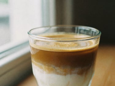 Close-up of a cortado in a clear glass, showing distinct layers of espresso and milk, soft side-lit window daylight, rich espresso and cream tones, shallow depth of field, 35mm film