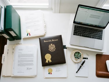 Overhead flat-lay of organized legal documents, university degree with a gold seal, and a laptop on a clean white desk, bright natural daylight, 35mm
