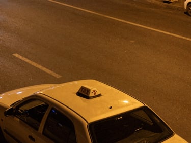 Taxi parked on an Elazığ street at night, amber street lamp light reflected on the vehicle roof, quiet empty street scene, wide shot, documentary style, no passengers visible