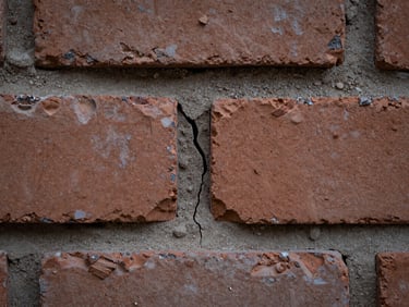 Macro close-up of a crumbling mortar joint between old clay bricks, cool diffuse light from above, fine sand particles and a hairline crack running vertically through the joint, no subjects staged