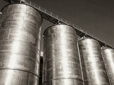 Low-angle heroic perspective of massive galvanized steel grain silos under dramatic side-lighting, high contrast black and white tones