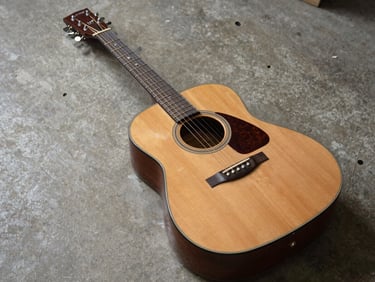 Flat overhead shot of a sunburst dreadnought acoustic guitar on a concrete workshop surface, natural north-facing daylight, no shadows
