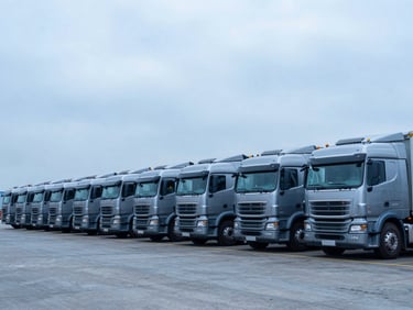 Wide-angle shot of heavy commercial trucks lined up at a maritime logistics terminal, metallic tones, overcast daylight, sharp focus.