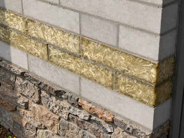 Overhead close-up of a wall section sample at a Dream Home Foundation site — layered cross-section showing insulation batt, concrete block, and external stone cladding, each material clearly differentiated, natural diffused daylight, no labels