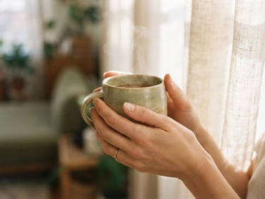 Soft focus close-up of hands gently holding a warm ceramic mug, natural light filtering through a sheer curtain in the background, muted sage and ecru tones, shallow depth of field, warm editorial photography.