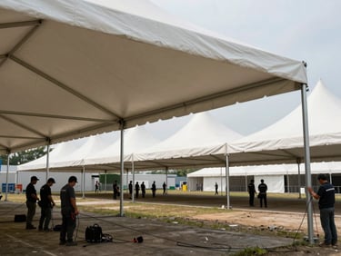 Wide shot of large white event tents erected on an open outdoor venue, aluminum poles and tensioned fabric visible, workers adjusting guy wires in the foreground, natural daylight