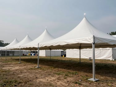 Wide shot of large white event tents staked on an open field, aluminum poles and tensioned fabric visible mid-installation, industrial overhead light, no crowds