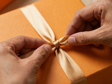 Macro close-up of hands tying a heavy gold silk ribbon around a textured saffron-colored box, warm editorial food photography, soft shadows