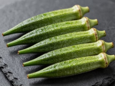 Fresh bright-green okra pods arranged in parallel on a dark charcoal slate surface, natural side-lighting highlighting ribbed texture, true colour, no filters