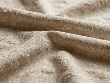 Close-up of heavy raw Belgian linen fabric, highlighting the rich organic texture and warm oatmeal tone under soft side light.