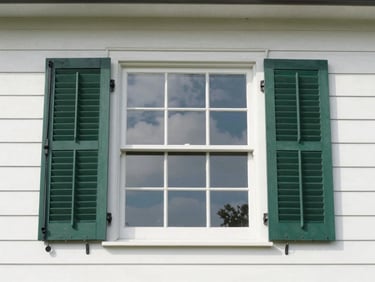 Exterior shot looking up at a clean two-story home window in daylight, sky reflected in the glass, cedar-green shutters flanking the frame