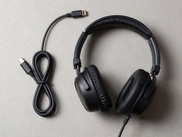 Overhead flat-lay of over-ear stereo headset with USB-C and 3.5mm cables coiled beside it on a neutral surface, studio strobe lighting, ear cushions and connector detail sharp, clean shadow