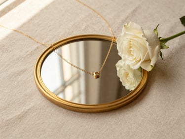 An editorial flat-lay of a gold-rimmed mirror, a delicate gold necklace, and a fresh white rose on a cream linen background, warm window reflections.