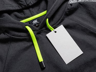 Flat-lay of a custom manufactured athletic hoodie, displaying a crisp woven neck label with custom branding, heavy-duty neon volt drawstrings, and a thick textured paper hangtag, dark industrial concrete background