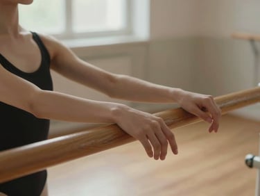 Close detail, soft indoor natural light — a ballet student's hands resting on the barre during a pause in class, warm light from a window, wooden floor visible below, the quietness of concentration in the frame