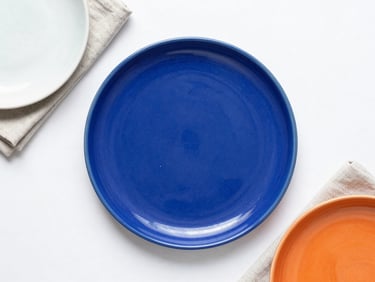 Flat lay of modern ceramic plates and linen napkins on a clean white background, vibrant cobalt accent, high-key lighting