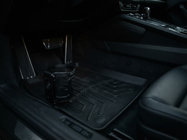 Low-angle close-up shot of an Apex Mounts cup holder securely clipped to a heavy-duty black floor mat in a Porsche 911 footwell, moody directional cabin light, deep shadows, matte textures, sharp focus, 50mm lens. The footwell is clean and minimalist, showing pedal box and part of the transmission tunnel.