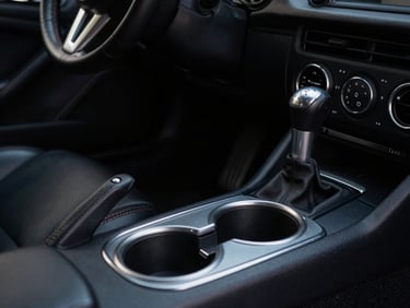 Close-up of Apex Mounts cup holder installed low in a Mazda MX-5 Miata footwell, showing ample clearance around the manual shifter, moody cabin light, deep shadows, matte textures.
