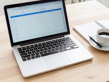A clean flat-lay of a modern laptop showing a spreadsheet, a notebook with a pen, and a coffee cup on a light wooden desk, bright natural daylight, 35mm