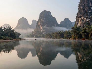 Panoramic view of Vang Vieng karst limestone mountains reflected in the Nam Song river at early morning, mist clinging to the base of peaks, a bamboo raft visible in the mid-distance, soft diffused daylight