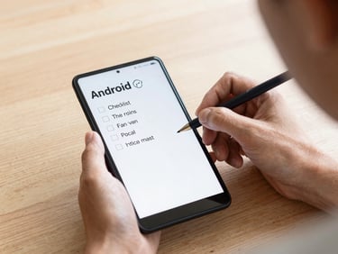 Over-the-shoulder shot of a hand writing a plain-text checklist on an Android device, soft overcast daylight, wooden table background.