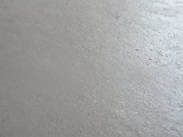 Vista macro de superficie de microcemento curado mate, textura de llana fina bajo luz rasante lateral extrema, tonos gris cemento y titanio, composición minimalista e industrial.