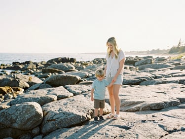 Hallie and her son laughing while exploring a rocky Maine beach, medium-wide environmental shot, warm lifestyle photography, soft morning sun.