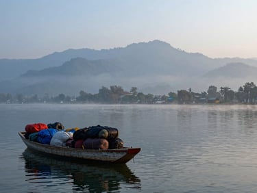 Wide panoramic frame of Dal Lake in Kashmir at dawn, a shikhara boat being loaded with luggage in the left foreground, misty mountains in the background, cool blue-grey morning light