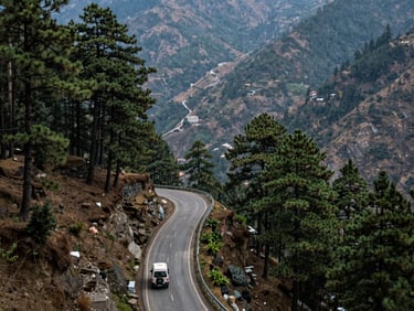 Wide shot of a mountain road in Uttarakhand with a vehicle winding through dense pine forest, natural daylight, valley visible below