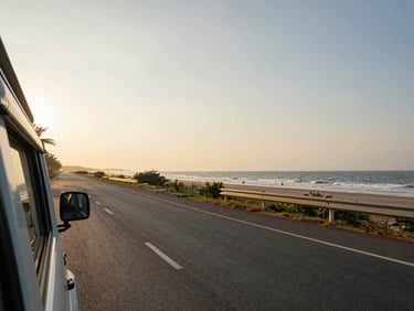 Wide documentary shot of a Tamil Nadu coastal road with a vehicle in the frame, Bay of Bengal visible to one side, golden-hour directional light