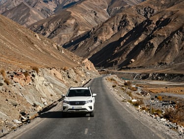 Wide environmental shot of Spiti Valley highway in Himachal Pradesh, a white SUV on a narrow mountain road, barren ochre hills flanking both sides, harsh midday light casting sharp shadows
