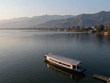 Wide environmental shot of Dal Lake in Kashmir from a high vantage point, a houseboat visible at the water's edge, mountains in background, morning light