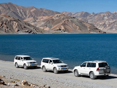 Panoramic wide frame of Pangong Lake in Leh-Ladakh, a convoy of two white SUVs on the lakeside gravel road, deep blue water and barren mountains behind, bright high-altitude daylight
