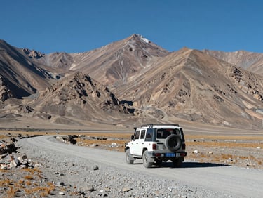 Wide shot of Leh-Ladakh high-altitude plateau with a 4x4 vehicle on a gravel road, barren mountains in the background, sharp midday sunlight