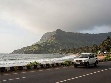 Wide environmental shot of a coastal Maharashtra road with a vehicle in the foreground, the Western Ghats visible in the background, overcast morning light