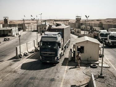 Wide-angle environmental shot of a heavy-duty transport truck stopped at a modern desert border checkpoint in Iraq, harsh high-contrast daylight, desaturated industrial tones, cinematic grit, no text.