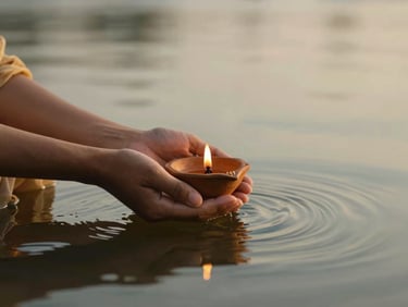 Dignified hands gently holding a small lit clay lamp floating on calm river water, warm golden hour light, peaceful spiritual transition, serene editorial photography