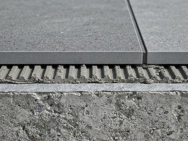 Wide-angle architectural photography, extreme macro cross-section of a high-performance commercial tiling system showing substrate, adhesive, and porcelain layers, cool gray concrete tones, sharp daylight, high contrast, 35mm lens
