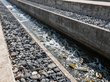 Wide shot of a concrete canal embankment reinforced with steel-mesh gabions filled with heavy stone, water flowing rapidly, bright daylight.