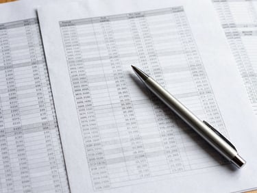Tight overhead shot of printed financial documents spread on a desk, columns of figures, a pen resting across the page, bright even studio light, no faces visible