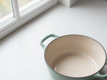 Overhead flat lay of a sage green cast iron dutch oven on a pristine white counter, soft north-facing window light, clean minimalist framing.