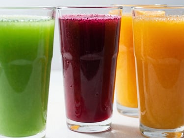 Three tall glasses of brightly colored cold-pressed juices—electric green, deep beet red, and vibrant orange—catching the sunlight on a clean surface.