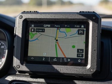 Extreme close-up of a ruggedized GPS tracking display mounted on a truck dashboard — an active route map with a live position dot shown on the screen, hard direct daylight cutting through the windshield casting sharp shadows across the device bezel; framing tight on the screen face, route lines clearly visible, no driver face in frame