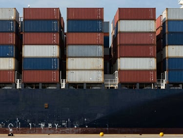 Wide shot of a container ship bow viewed from a low dock angle, stacked containers in regimented rows, hard midday sun overhead, deep shadows between container columns, no crew visible, geometric industrial composition