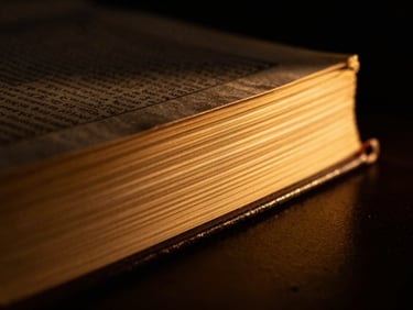 Chiaroscuro macro shot of gold-foil page edges of a closed sacred book, glowing amber light leak, dark background