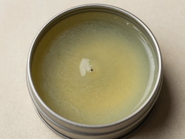 Extreme close-up overhead studio shot of a flat open tin of herbal lip balm on a neutral parchment surface, waxy surface texture and faint green-amber colour visible, bright studio daylight, no lifestyle props
